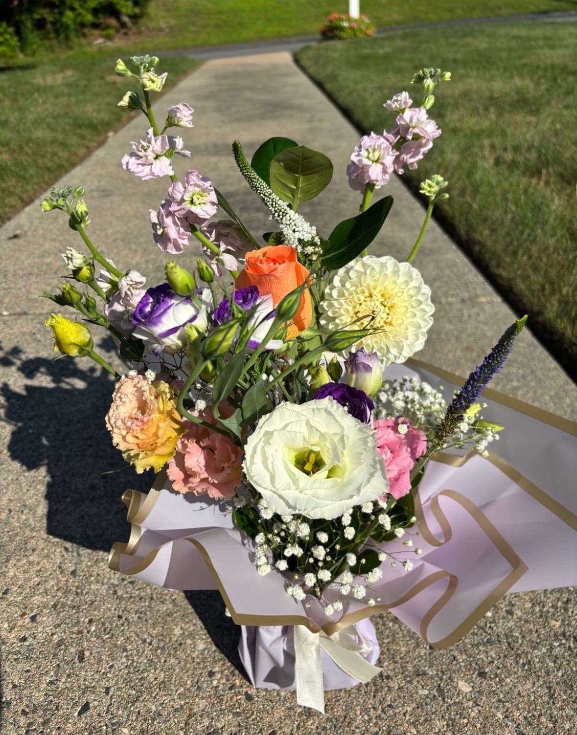 Wrapped arrangement of lilac stock, purple lisianthus, an orange rose and a cream dahlia on a sunny walkway